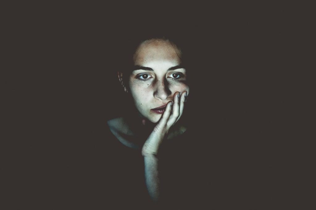 A person is seen gazing at a screen, illuminated by its light in an otherwise dark setting. They rest their face on one hand, looking contemplative with an expression of deep thought.