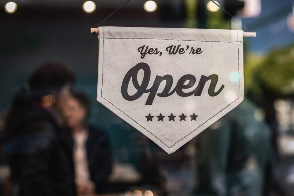 A white fabric sign hangs in a store window with the text "Yes, We're Open" and five stars below it. The background shows blurred silhouettes of people and lights.
