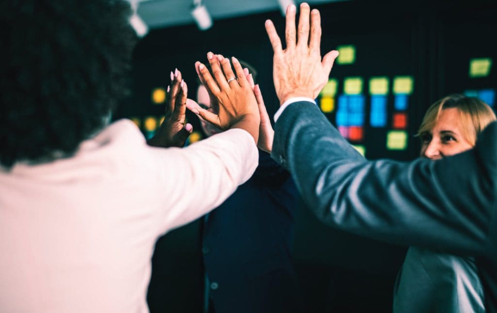 A diverse group of people wearing business attire are seen from the side giving a high-five to each other. The background is dark but features colorful sticky notes on a wall. The mood appears positive and energetic, suggesting teamwork and celebration.