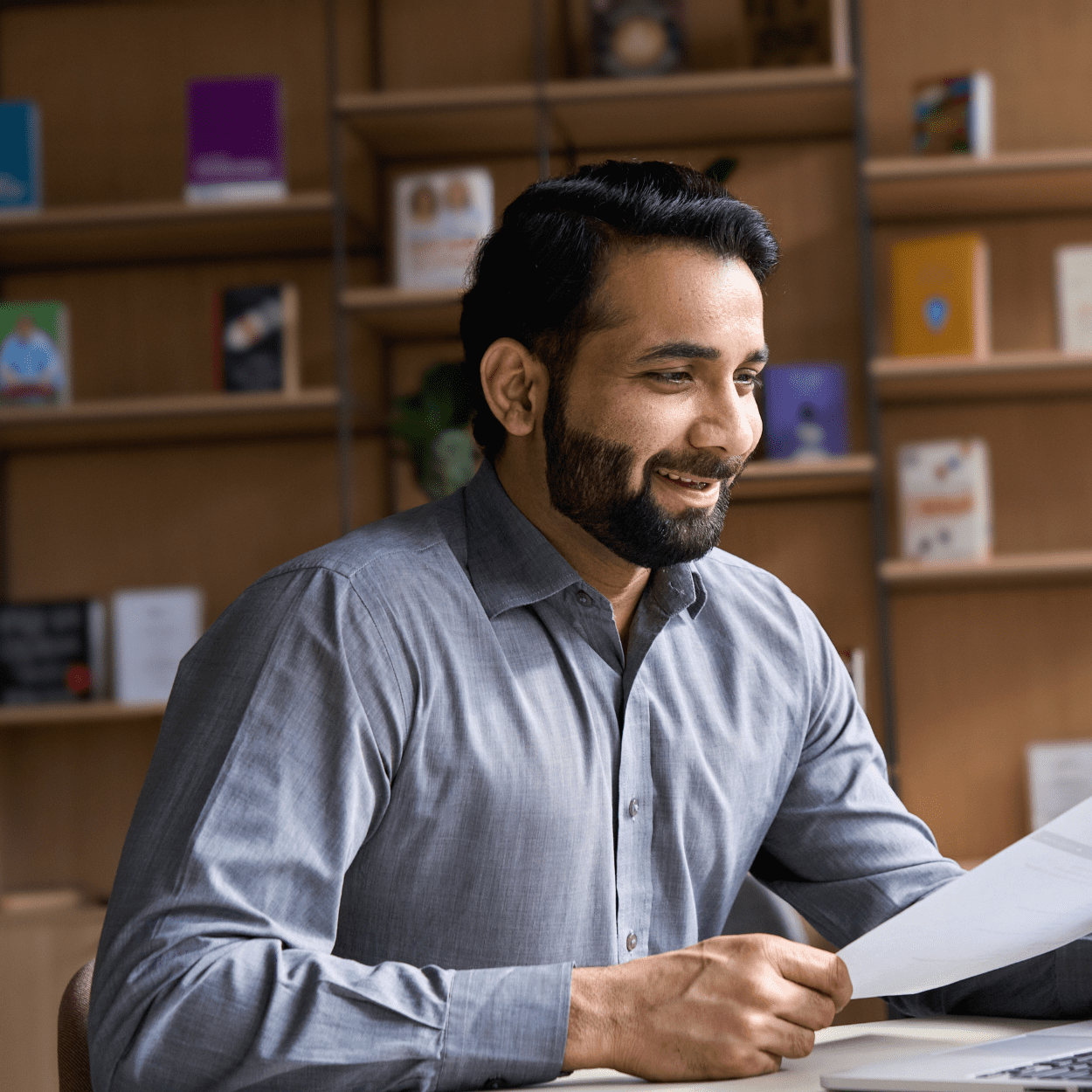 A man dressed in a grey button-up shirt sits at a desk in an office or library setting. He is smiling and holding a sheet of paper. Shelves with books and notebooks are visible in the background.